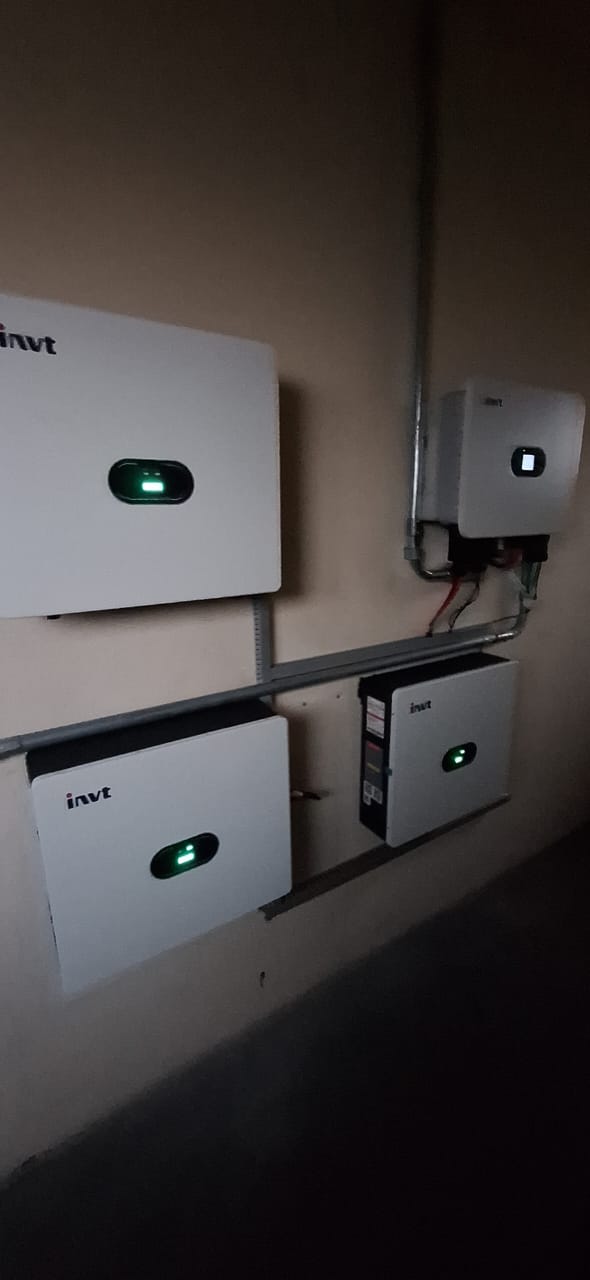 Equipamentos brancos de bateria e inversor fixados na parede de uma sala técnica, com luzes verdes de funcionamento acesas.