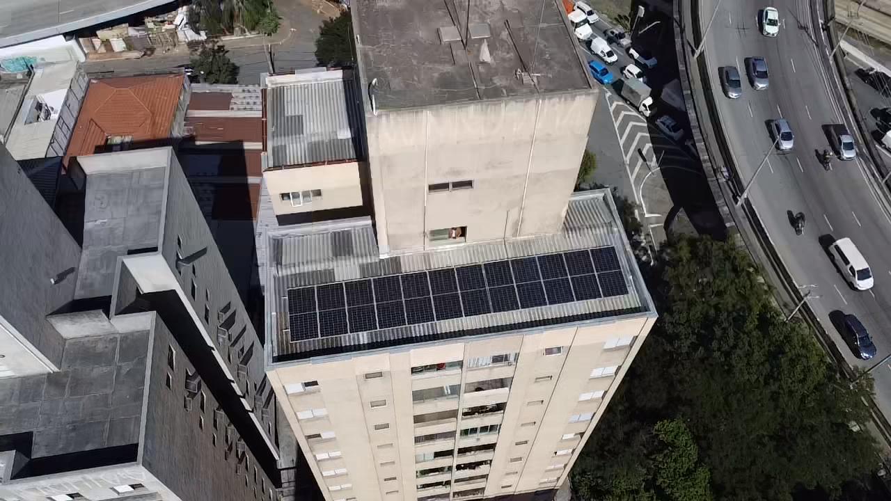 Vista aérea de um prédio residencial com uma fileira de painéis solares no telhado, ao lado de uma avenida.